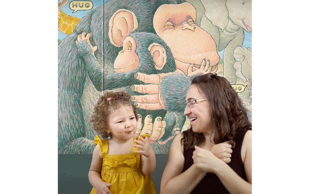 Mother and young daughter are signing together. A page from the book Hug is shown in the background, which shows a mother and baby chimp hugging.