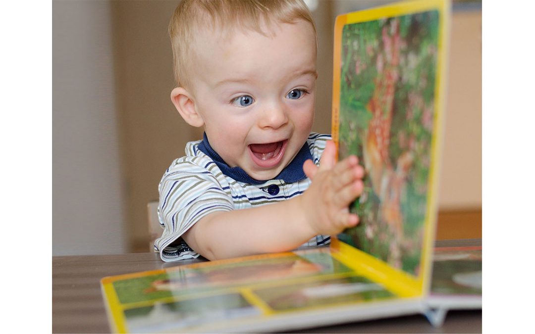 baby boy is holding up the page of a book. He has a big smile on his face and looks excited.